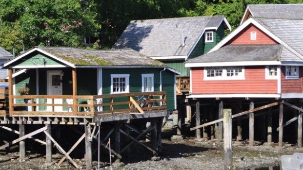 Two colorful stilt houses on a rocky shoreline, one green and one red, with a larger house behind them
