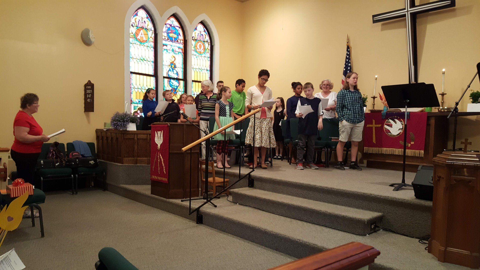 Choir singing from a stage in a church. Stained glass windows and a cross are visible.