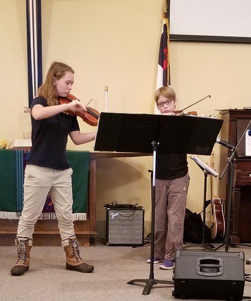 Two people playing violins on a stage, music stand in between.