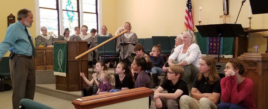 A man uses a long stick to point to people in a church setting with a group of children and other adults.