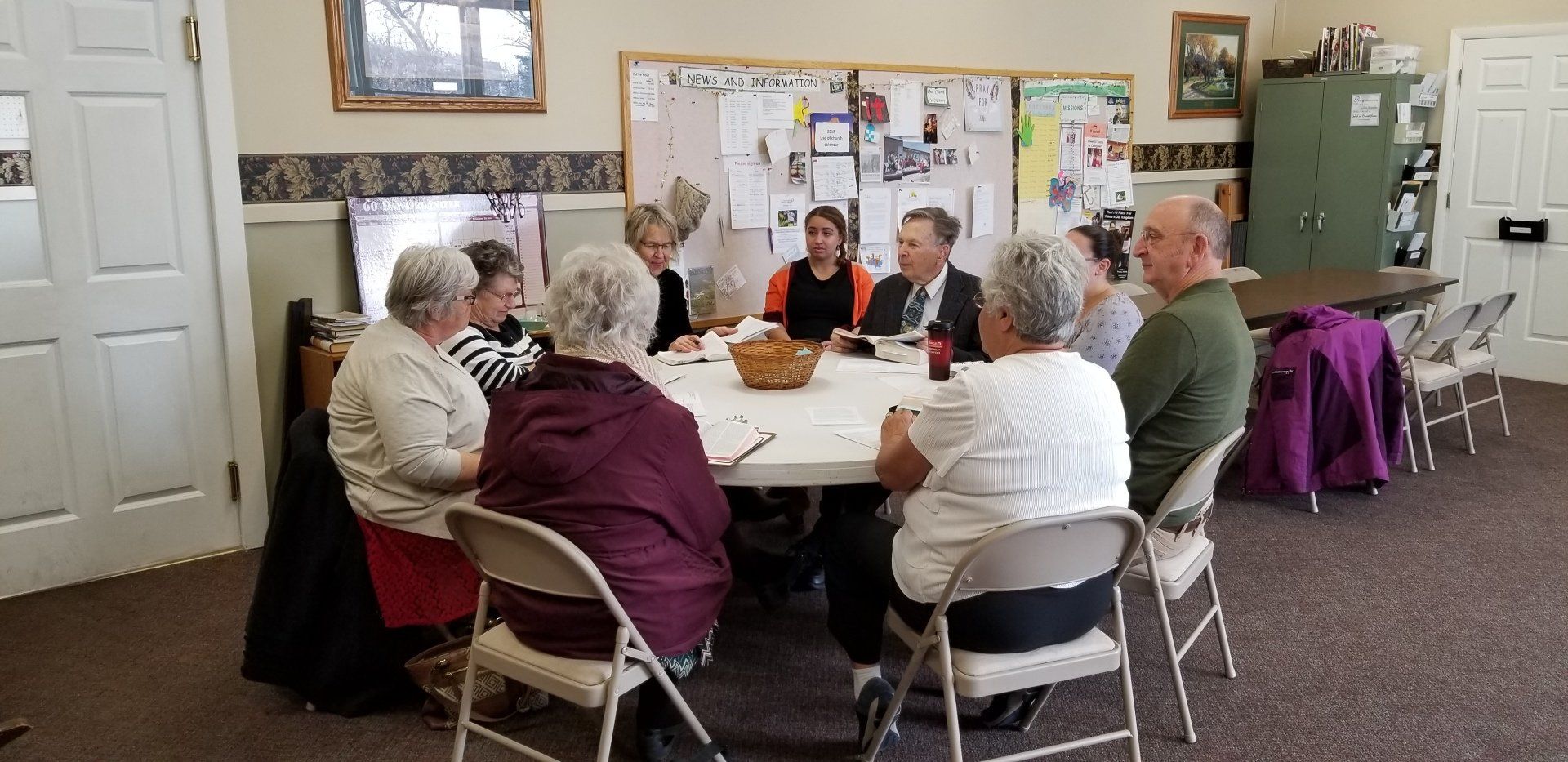 Group of people seated around a table, possibly at a meeting, in a room with a bulletin board and framed art.