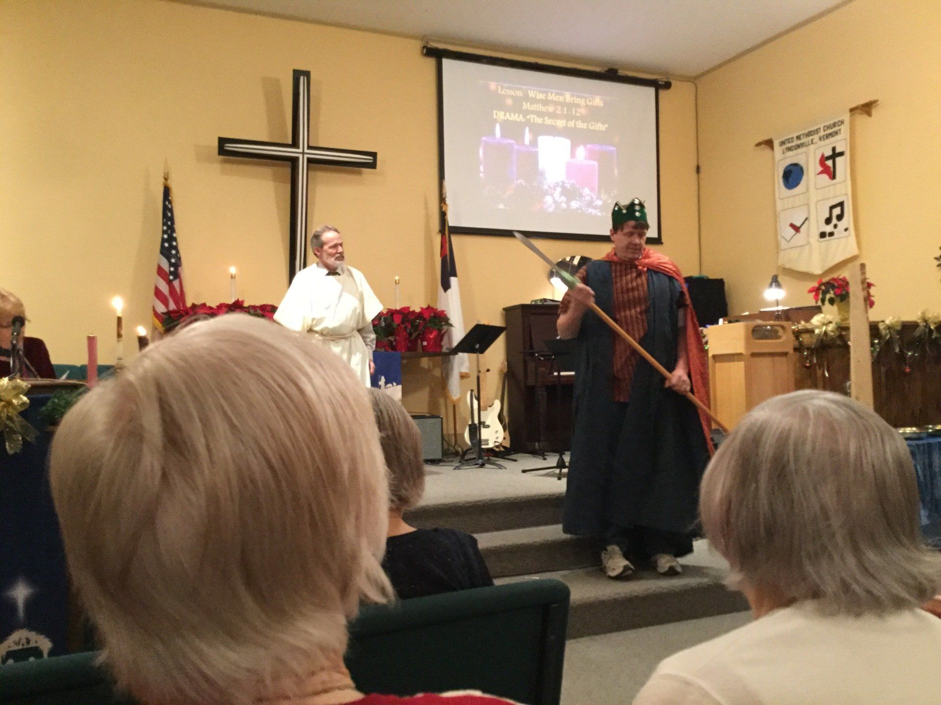 Man in costume holding staff, another in robe, on stage in a church, cross in background.