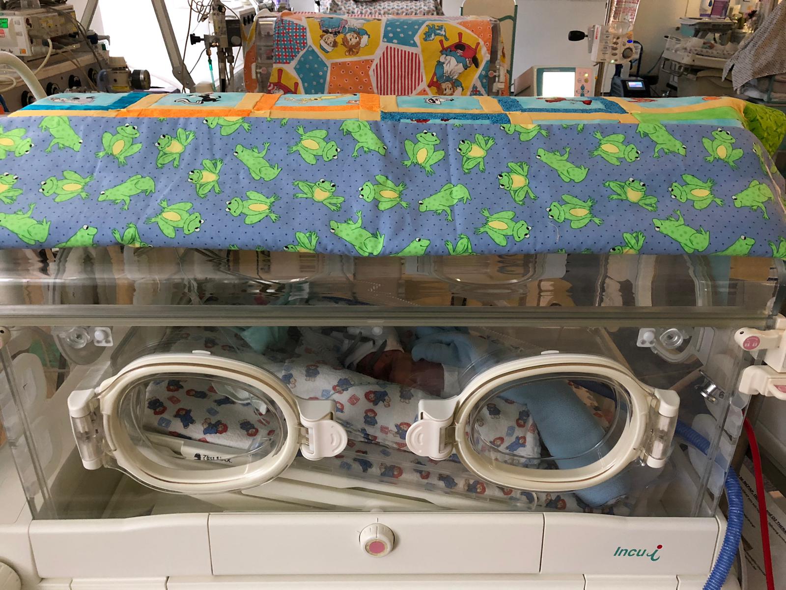 A baby in an incubator, covered with a blue blanket with frog patterns.