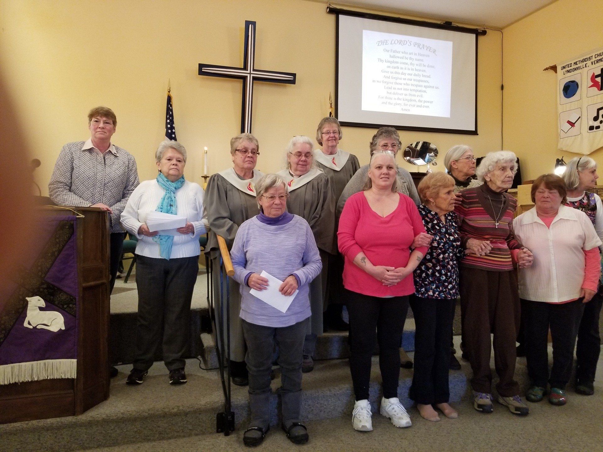 Choir stands on a church stage, in front of a cross and projection screen.