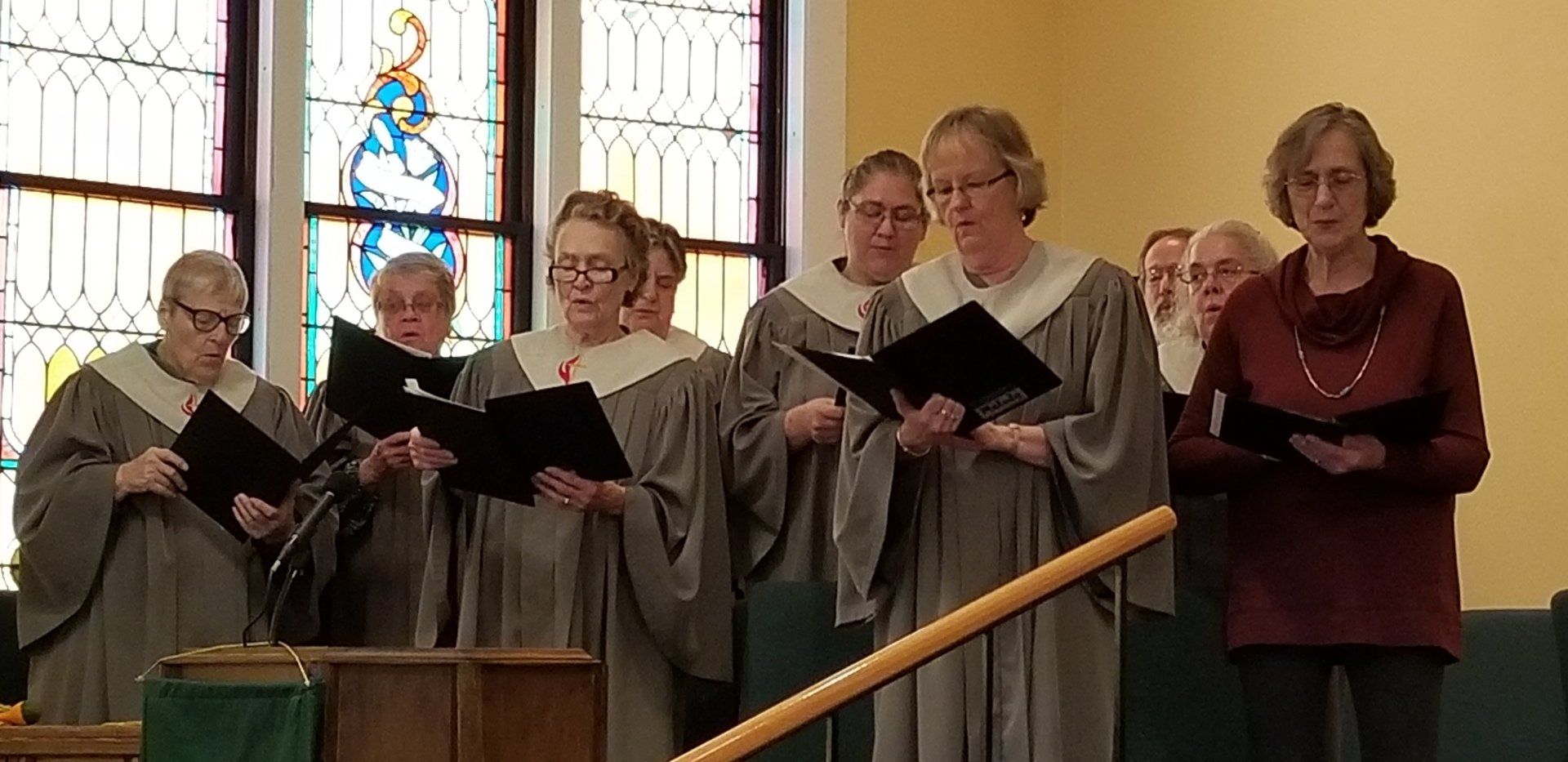 A choir in gray robes sing, holding songbooks, near a stained glass window in a church.
