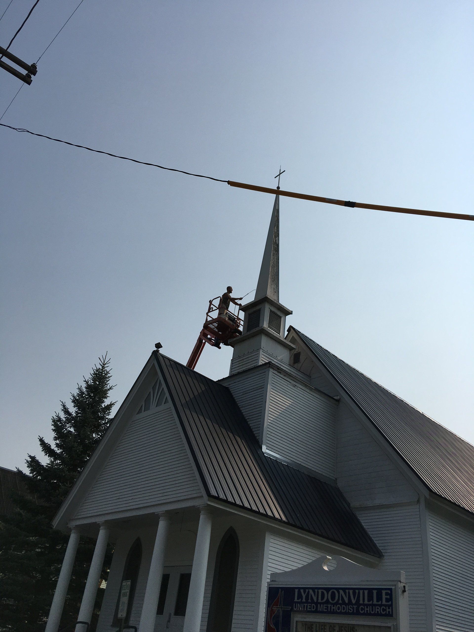 Worker in a lift repairs church steeple. White church with dark roof under a clear blue sky.
