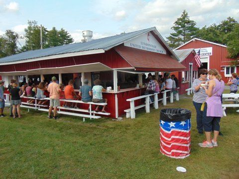 Red concession stand with people, outdoor seating, and an American flag trash can.
