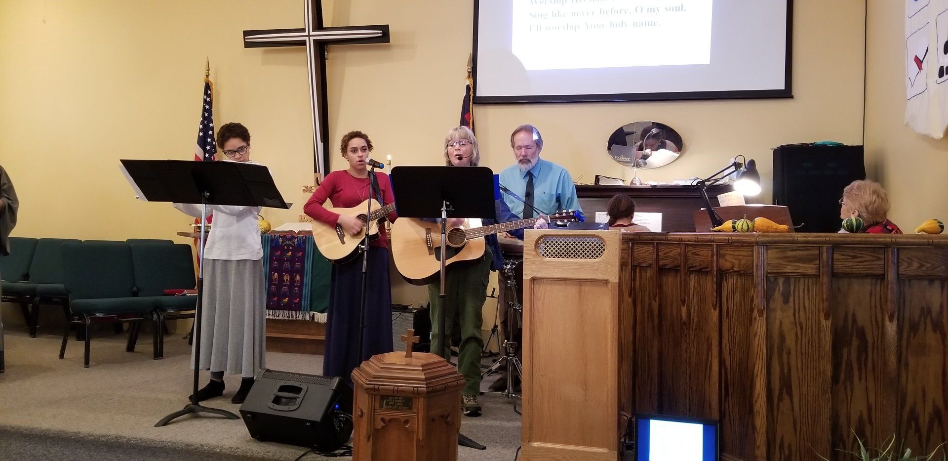A band performs in a church. Four people sing and play guitars. A cross hangs on the wall.
