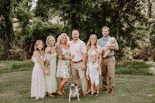 Family portrait outdoors, smiles, greenery, pug, dresses and button-up shirts, sunny day.