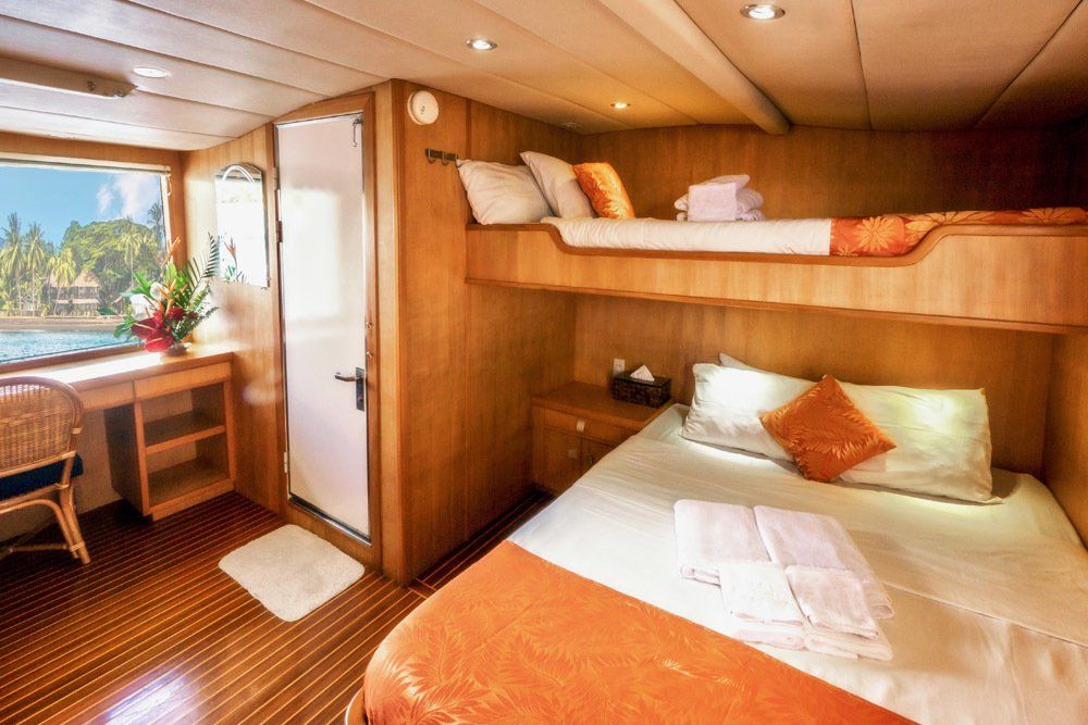 Cabin on a boat with bunk bed, desk near window, and bathroom door. Wooden interior, orange accents.