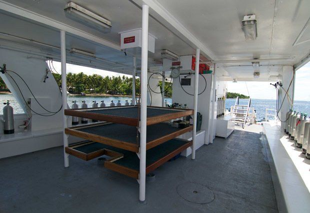 Deck of a dive boat with three-tiered equipment racks, tanks, and a view of the ocean.