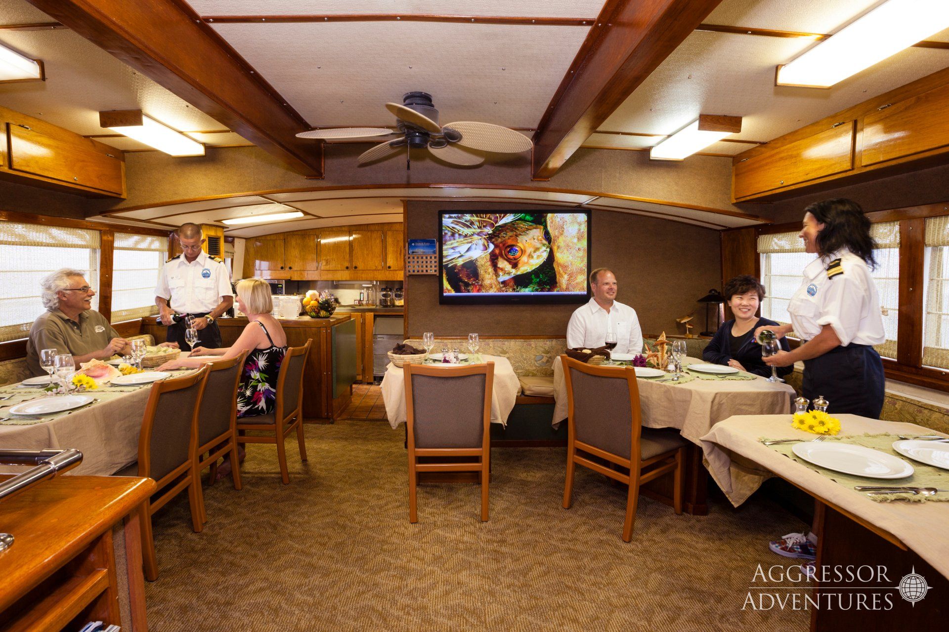 Dining room on a boat. Diners seated at tables, waiters serve them. Wooden ceiling, TV on wall.