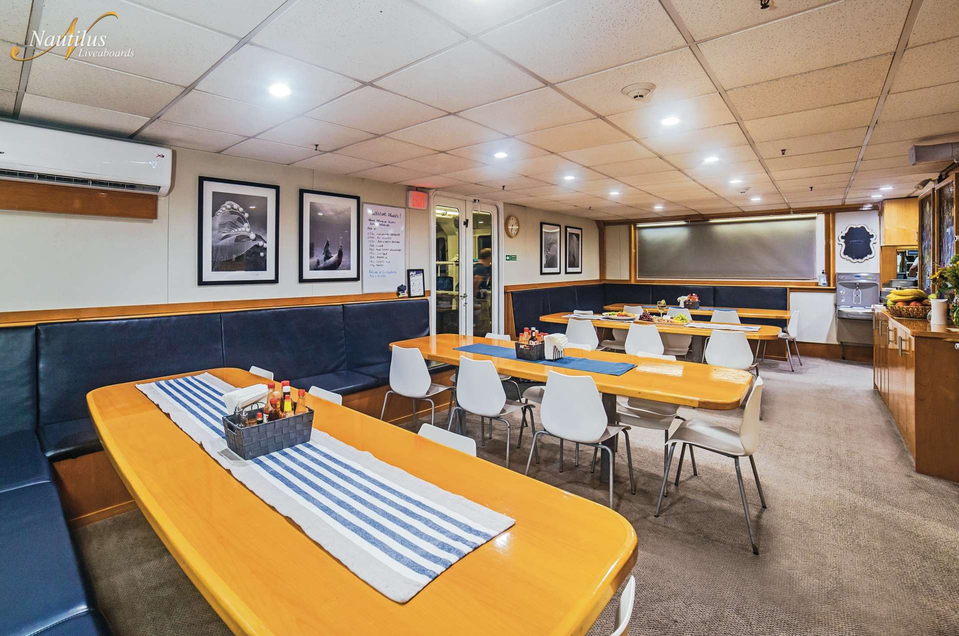 Dining area inside a ship with long tables, blue booth seating, and white chairs.