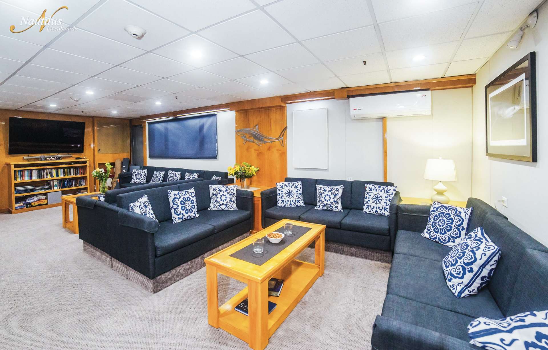 Lounge area with dark sofas, patterned pillows, a wooden coffee table, and a large TV on a boat or ship.
