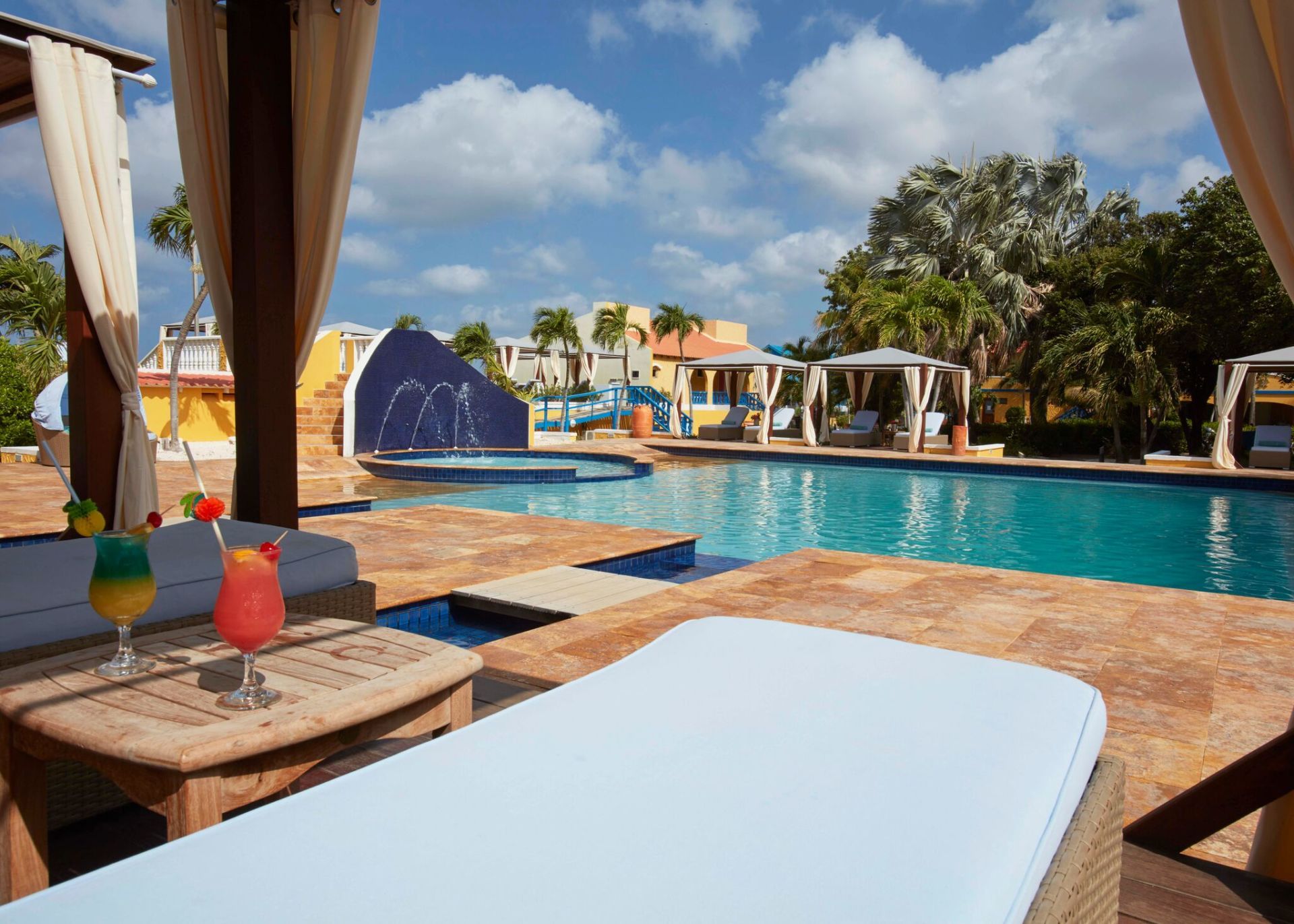 Poolside lounge area with cocktails, turquoise pool, and cabanas under a sunny sky.