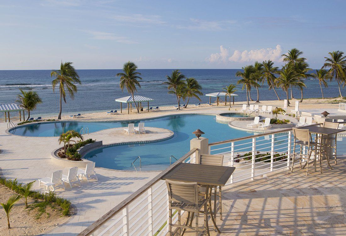 A resort pool area with a beachfront view, featuring palm trees, blue water, and white lounge chairs.