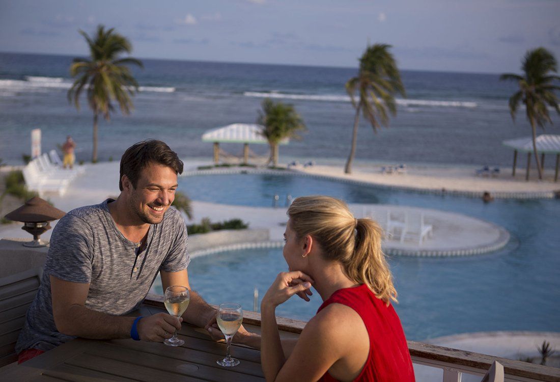 Couple enjoying drinks on a balcony overlooking a pool and ocean.