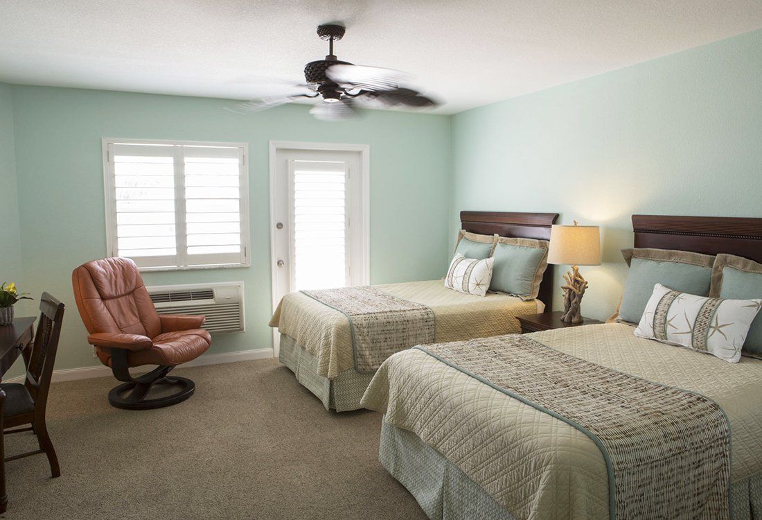 Bedroom with two beds, armchair, desk, and door to outside. Pastel green walls, tan carpet.