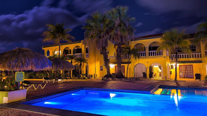 Yellow hotel at night with a pool, palm trees, and tiki hut.
