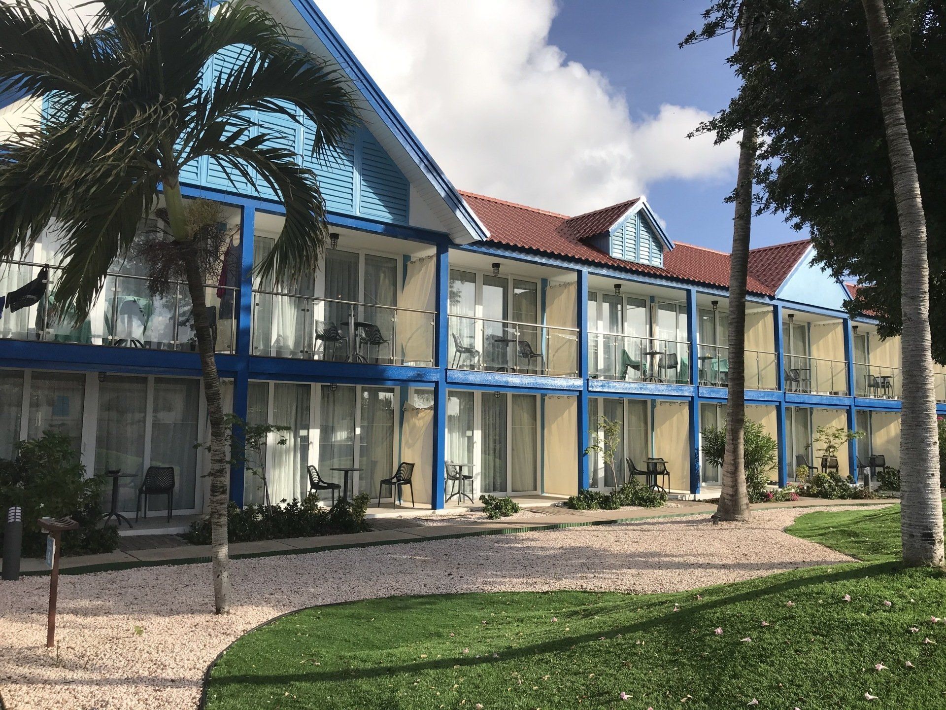 Two-story hotel building with blue trim, red roof, and balconies, on a grassy lawn with palm trees.