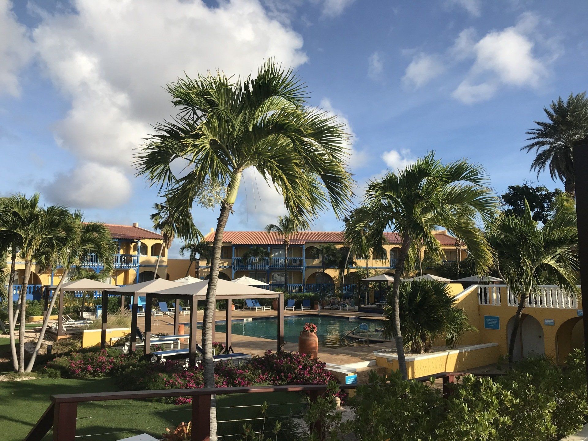 Tropical resort scene with pool, palm trees, and yellow buildings against a blue sky.