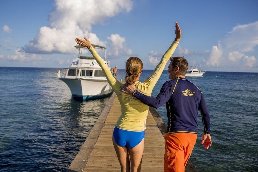 Couple on a pier with arms raised toward the ocean, boats in the background, sunny day.
