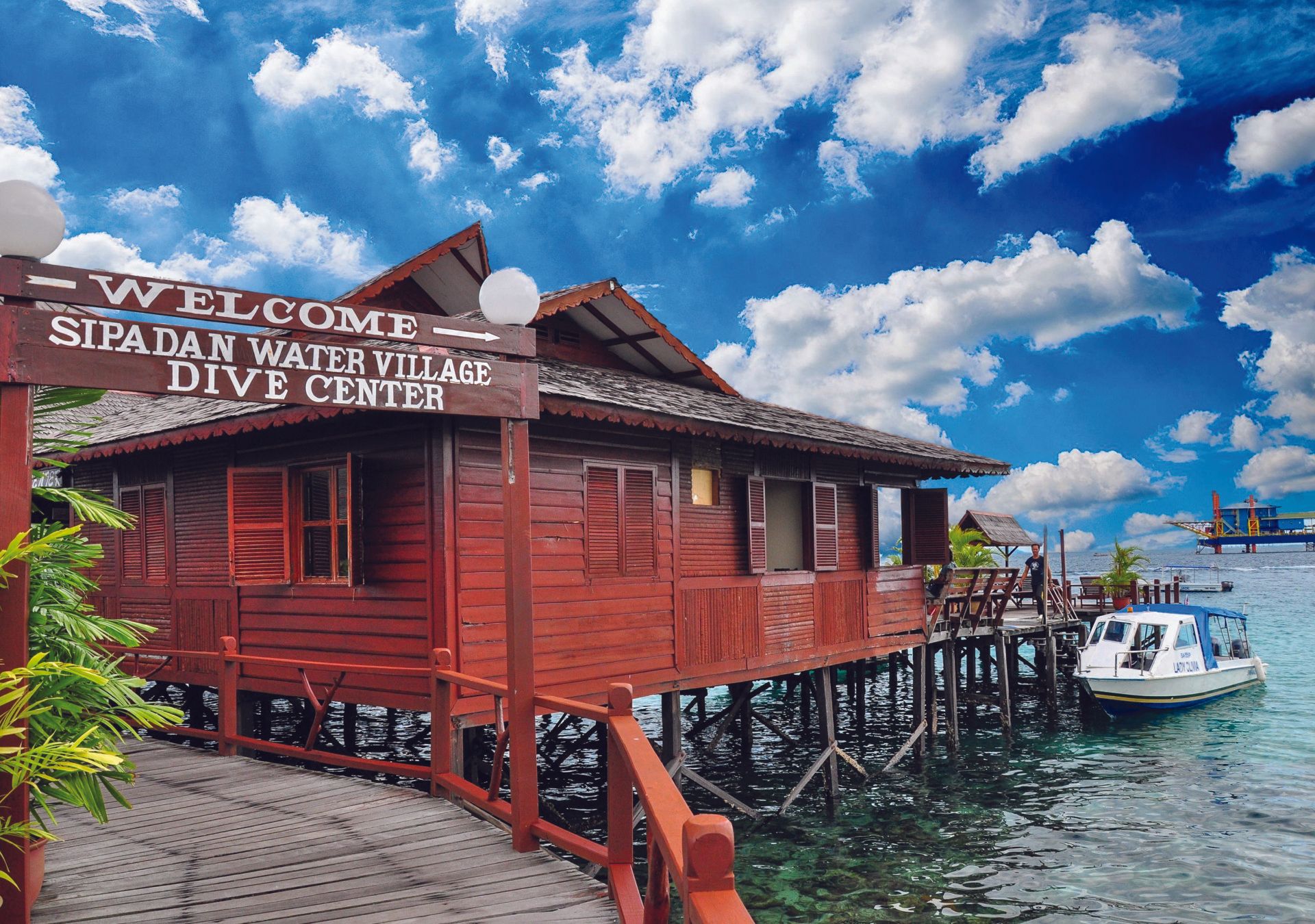 Wooden Sipadan Water Village Dive Center on stilts over turquoise water under a cloudy sky.