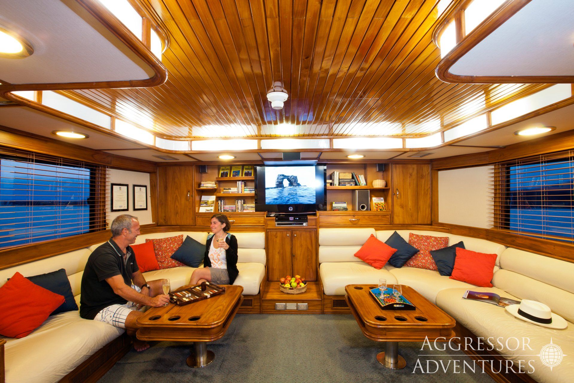 Interior of a boat lounge with a couple seated on a cream sofa, watching TV. Wooden paneling, blue sea visible outside.