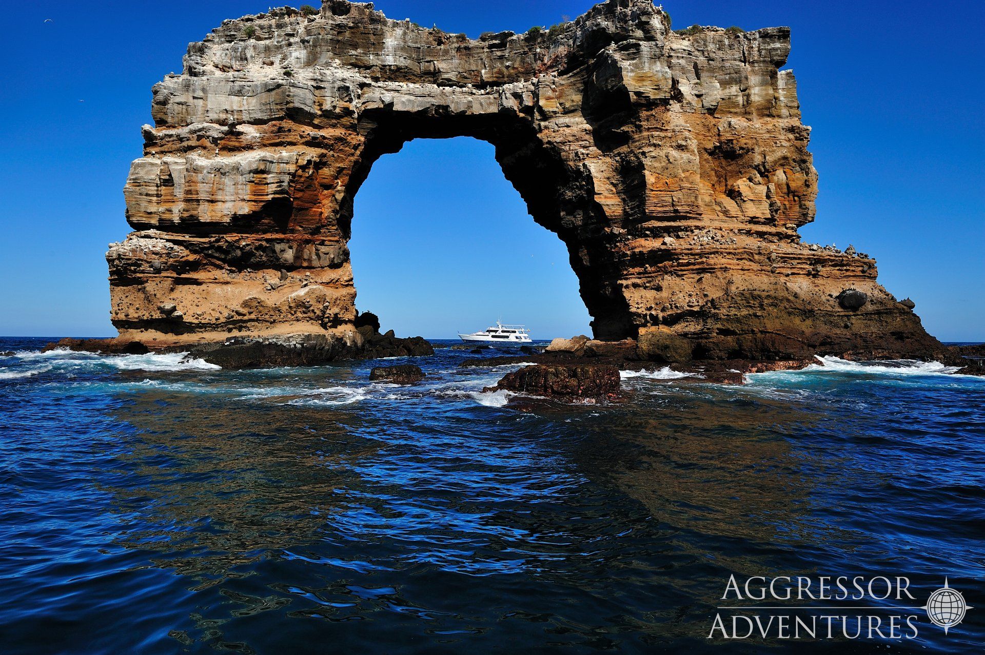 Rock arch in the ocean, blue water, boat in the distance, sunny day.