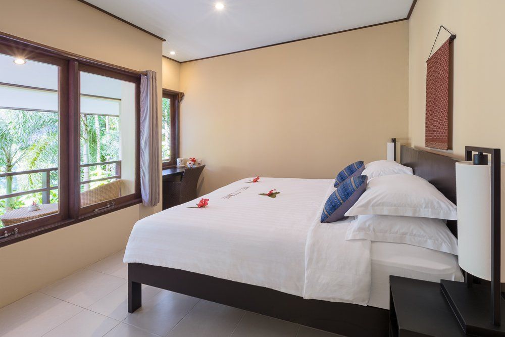 Bedroom with king bed, balcony view, beige walls, brown frame, white bedding, blue pillows.