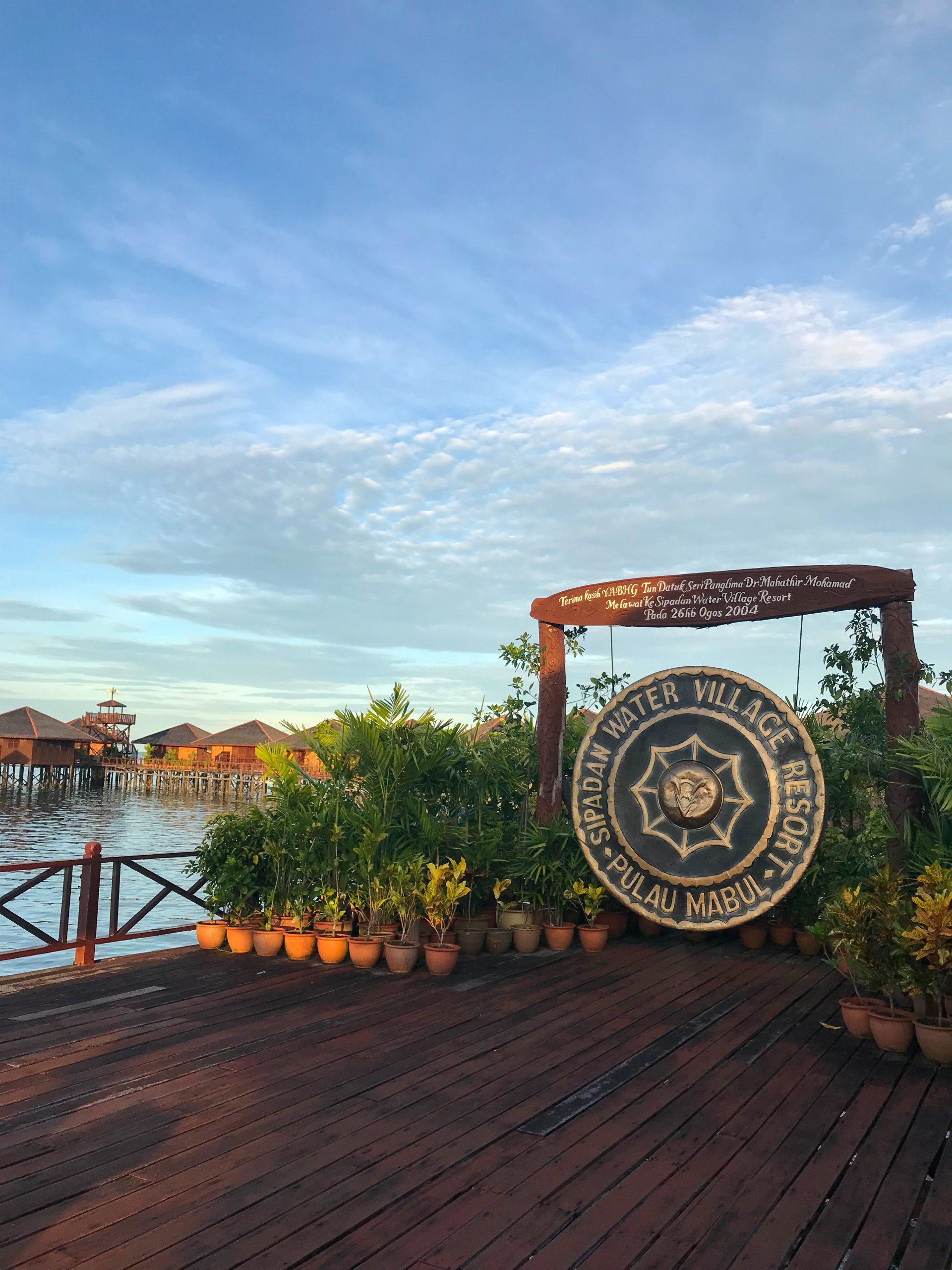 Wooden deck overlooking water with a large gong. Stilt houses in the distance. Blue sky with clouds.