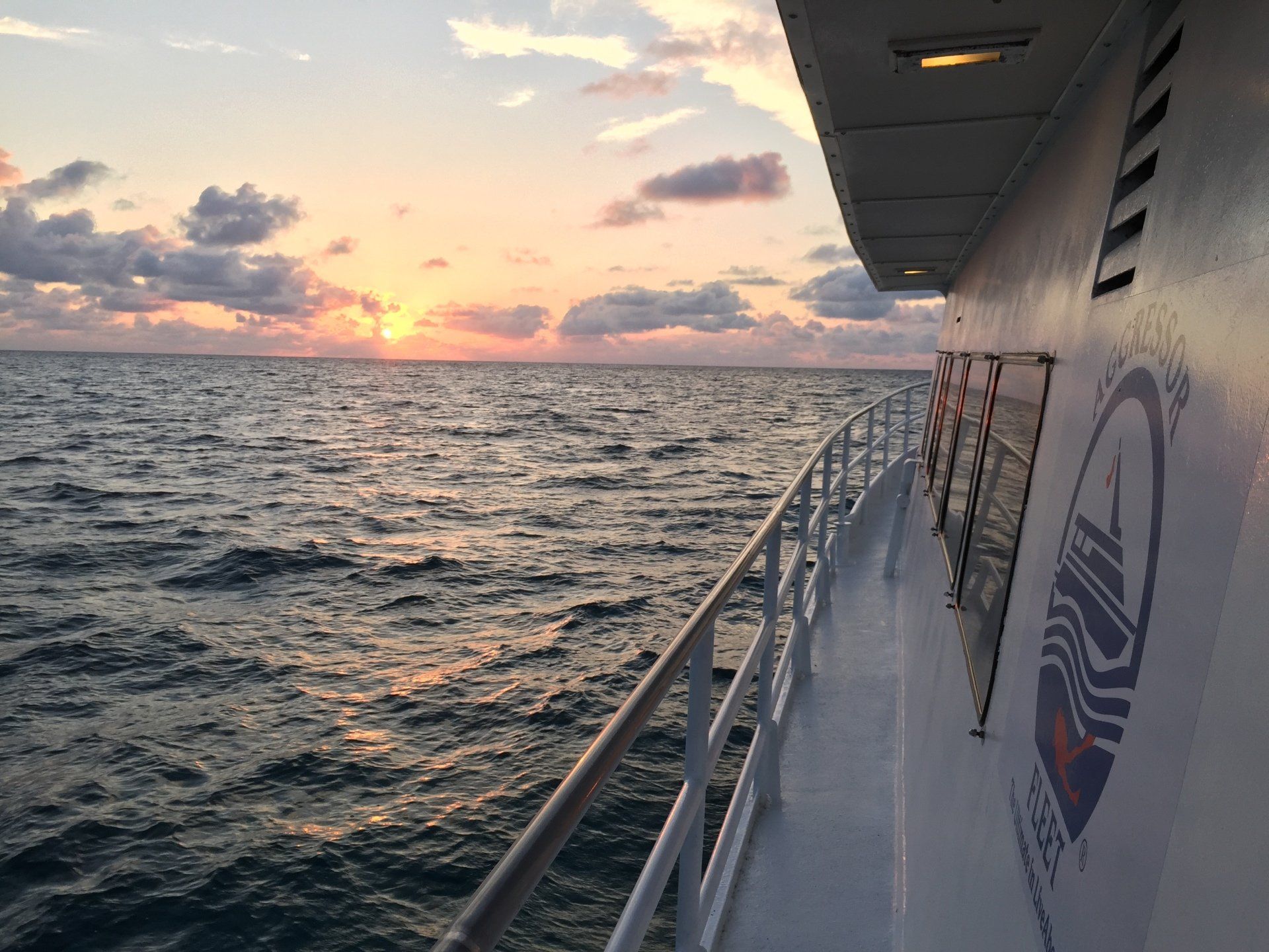 Sunset over ocean viewed from a boat's side, with railing, white hull, and logo.