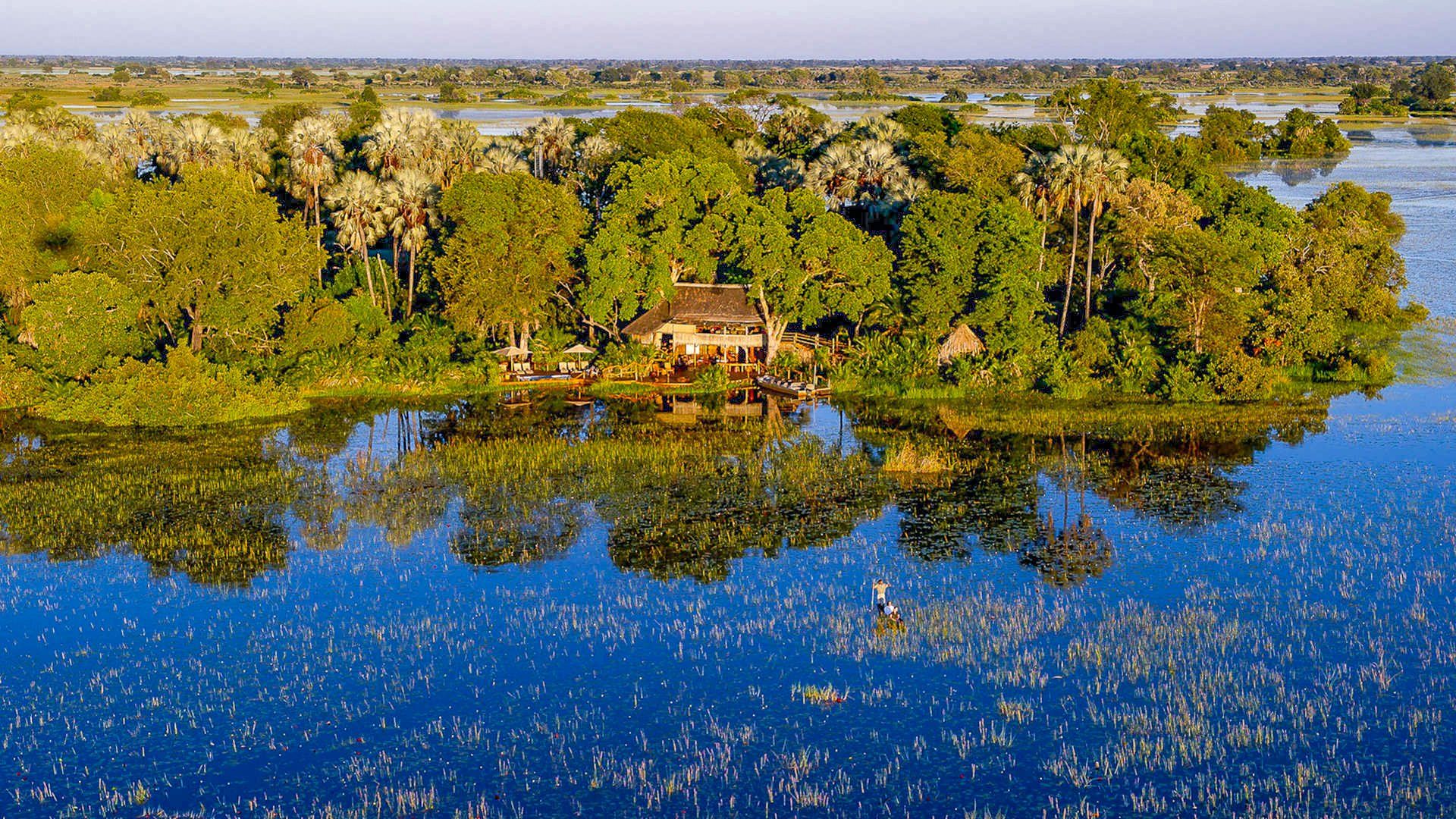 A lodge on a lush island surrounded by blue water. Trees reflect in the water's surface; expansive landscape in the background.