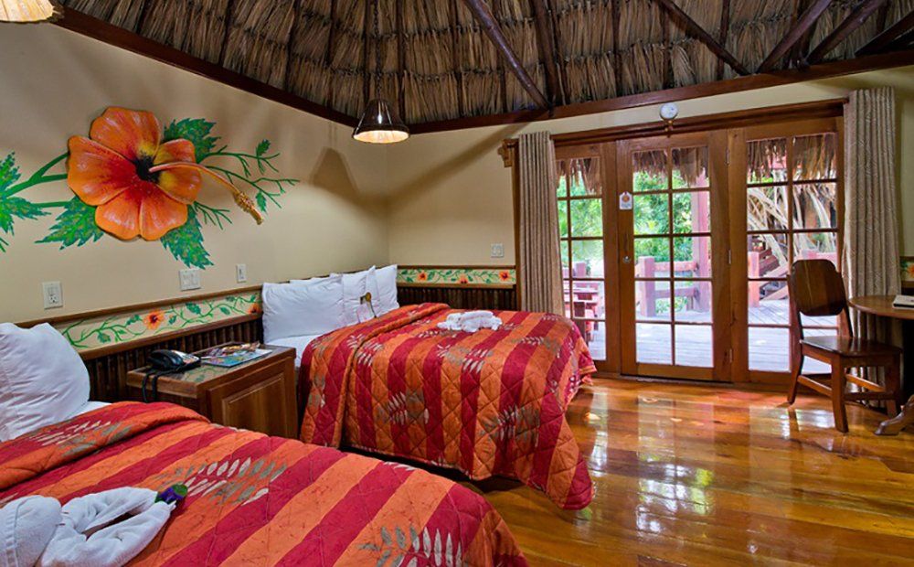 A hotel room with two beds, an orange floral quilt, and a view of an outdoor patio through open doors.
