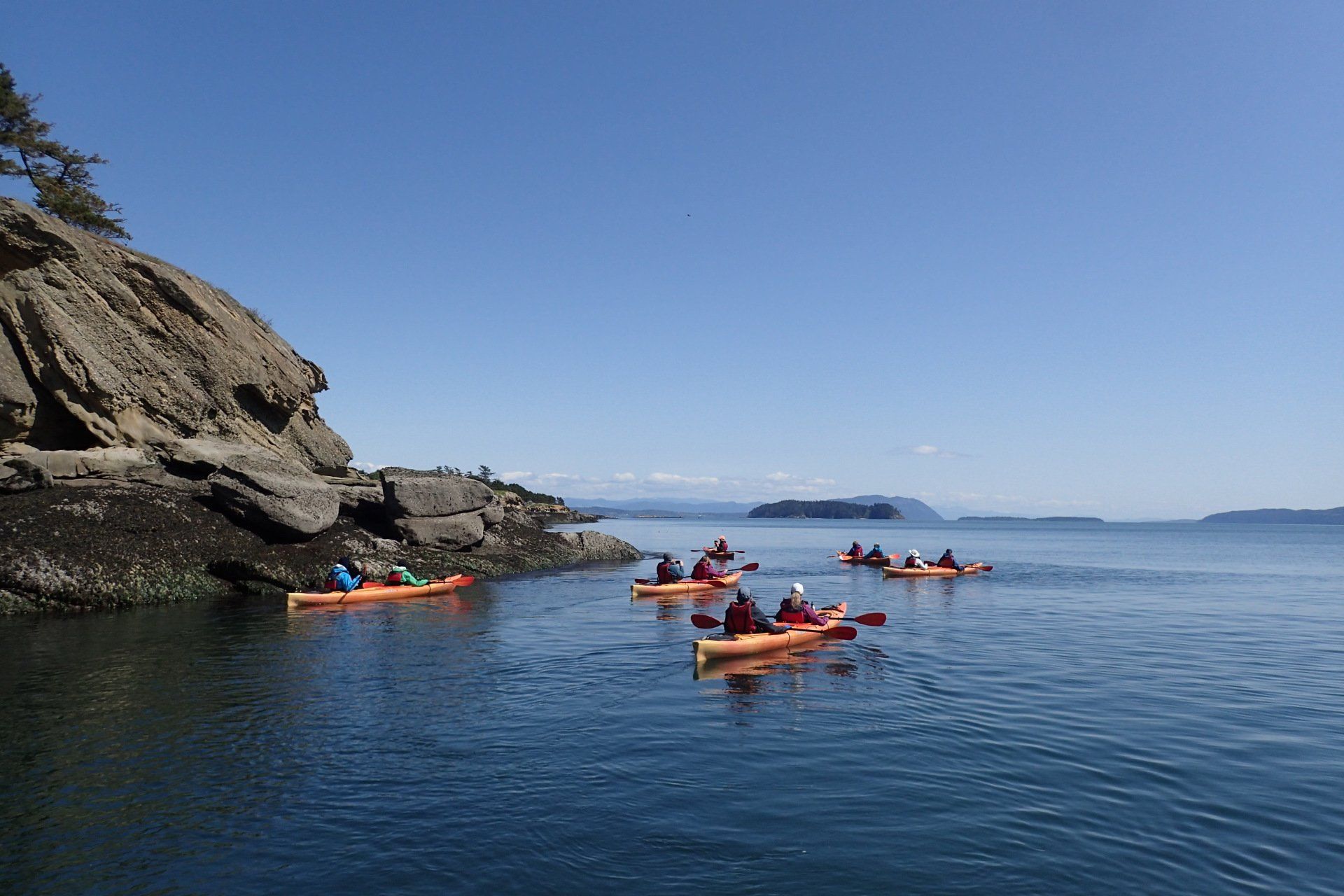 Kayakers paddle in orange kayaks near a rocky shore and islands under a blue sky.