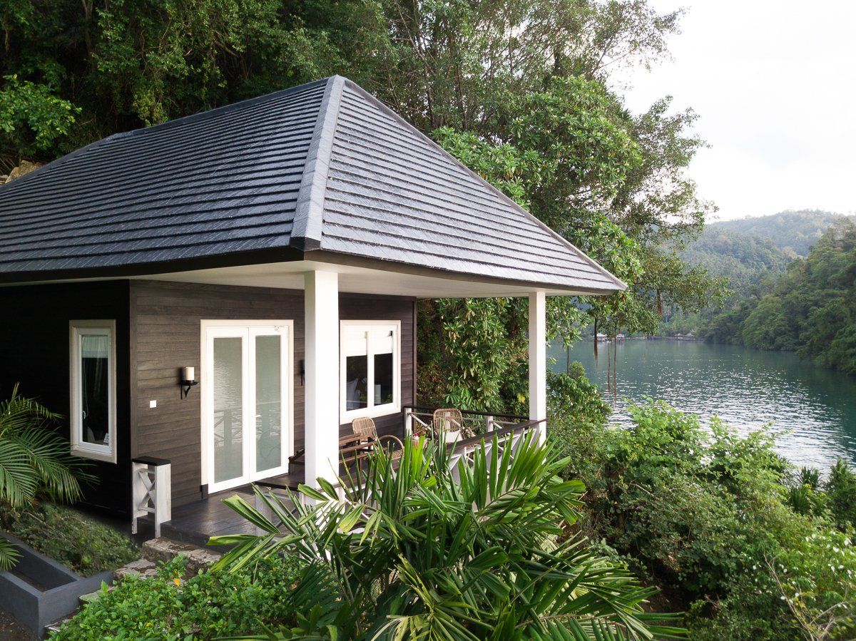 Cabin with dark roof overlooking a serene lake surrounded by lush green trees.