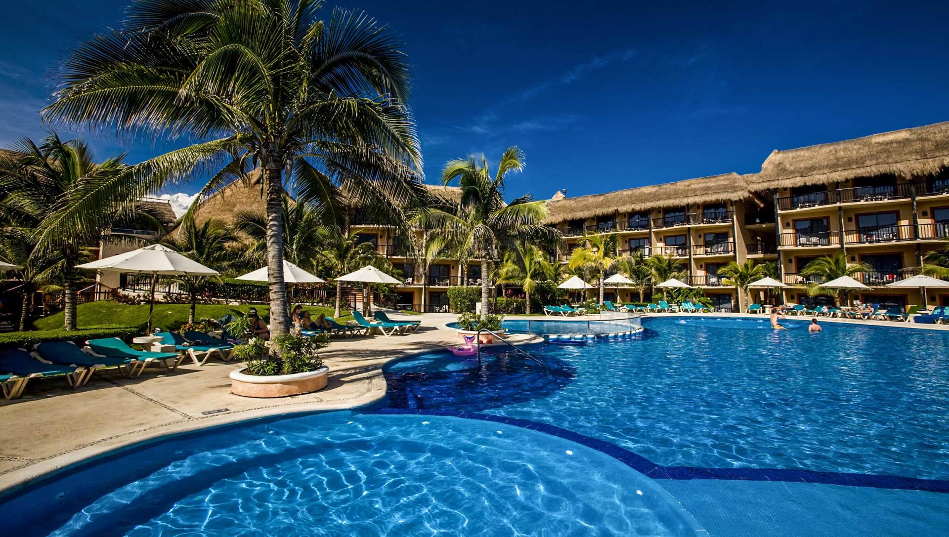 Poolside resort with palm trees, blue water, and thatched roof buildings under a bright blue sky.
