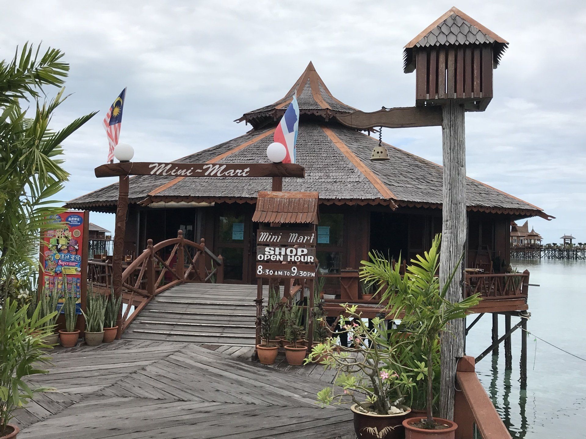 Wooden building on stilts over water with thatched roof, bridge, and Malaysian flags.