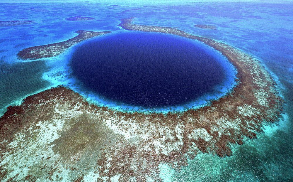 Aerial view of the Great Blue Hole in Belize, a dark blue circular sinkhole surrounded by coral reefs in clear turquoise water.