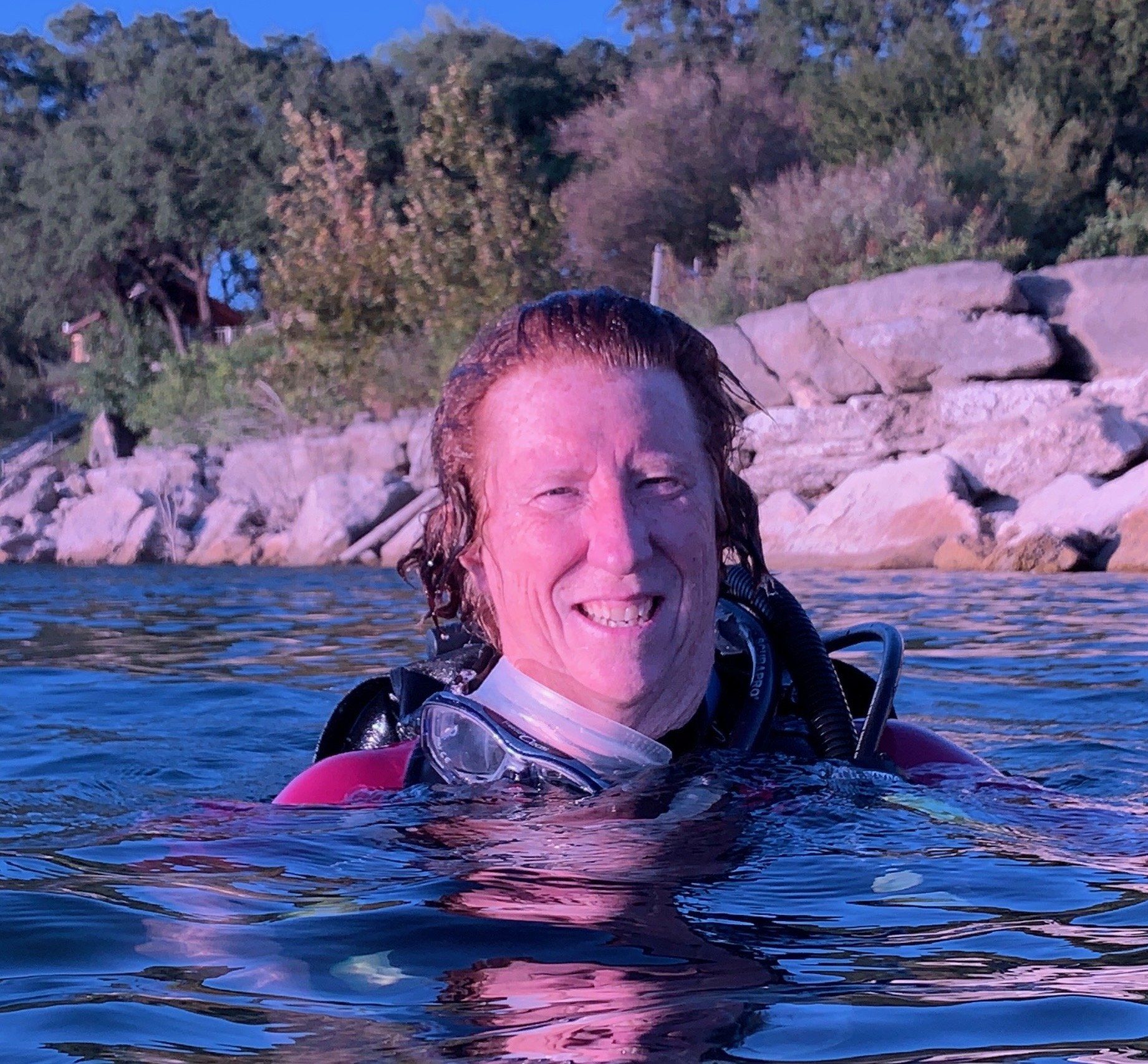 Diver in water with scuba gear, smiling. Trees and rocks in background.
