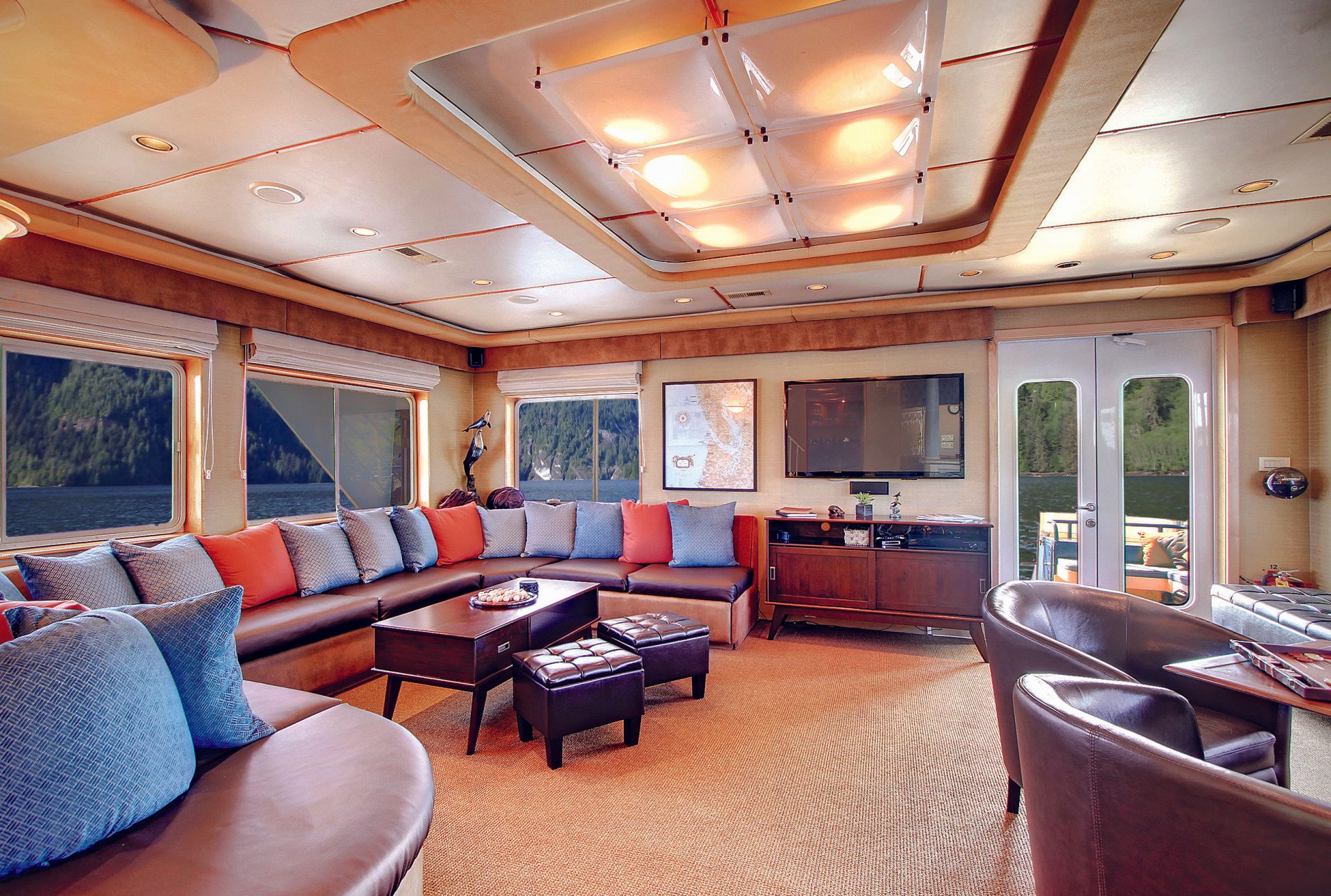 Lounge area on a yacht with a sectional sofa, coffee table, and large windows with views of a water and trees.