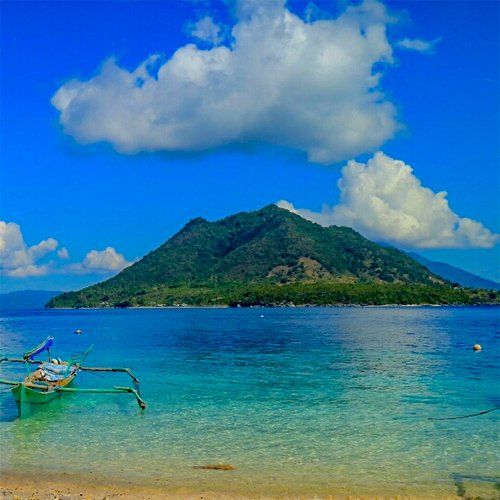 Blue ocean with clear shallows, small boat, green island under a bright blue sky with fluffy clouds.