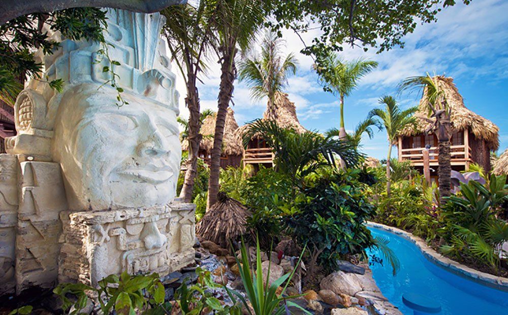 Stone Mayan-style sculpture, thatched-roof buildings, and blue waterway set amid lush tropical foliage.