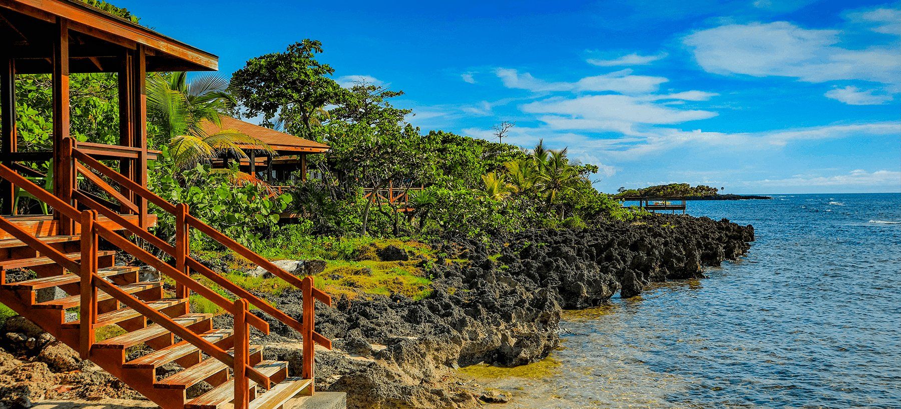 Wooden stairs and a gazebo overlooking a rocky coast and blue ocean under a bright sky.