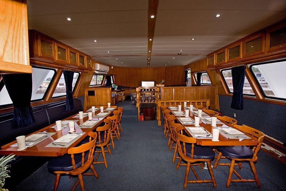 Interior of a boat dining area with tables set for a meal, blue carpet, wood paneling, and large windows.