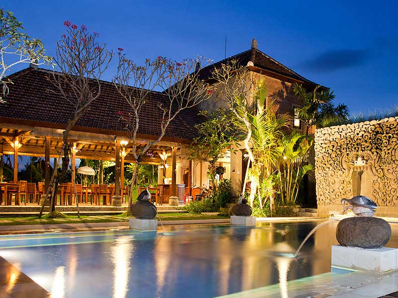 Nighttime shot of a lit pool with stone fountains and a traditional Balinese building in the background.