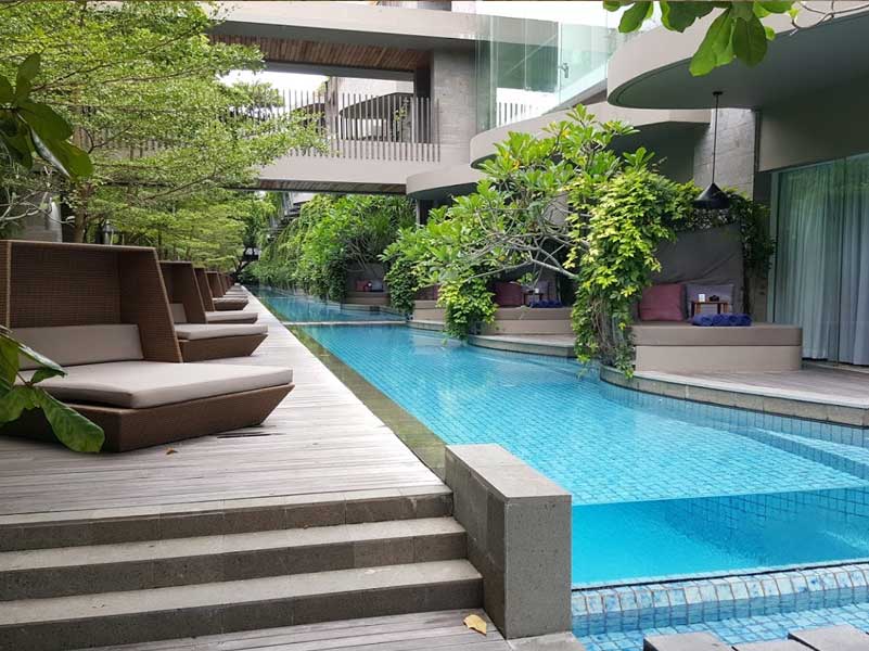 Pool with lounge chairs between hotel buildings, lush greenery, and blue water.