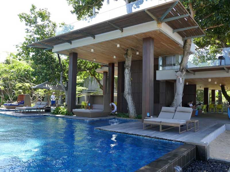 Poolside lounge area with wooden canopy, pool, and trees.