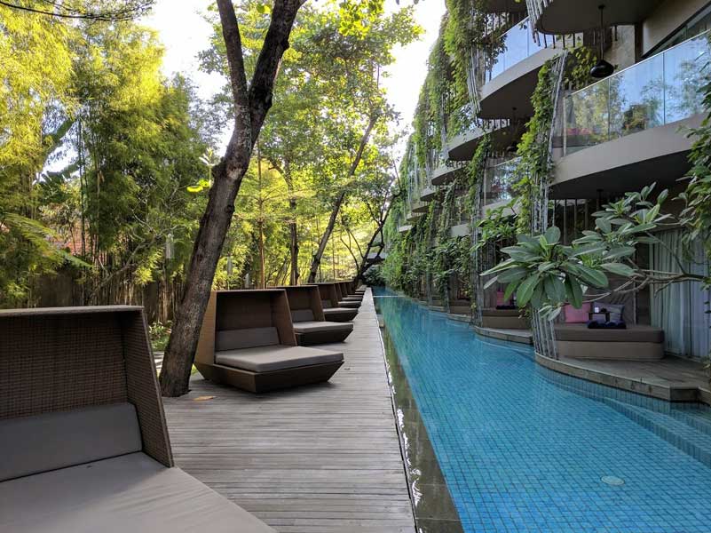 A long, narrow pool next to a row of lounge chairs and a building with balconies. Green trees surround.