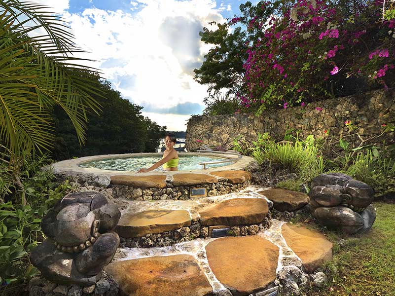 Person in a stone hot tub surrounded by lush greenery, with a cloudy sky in the background.